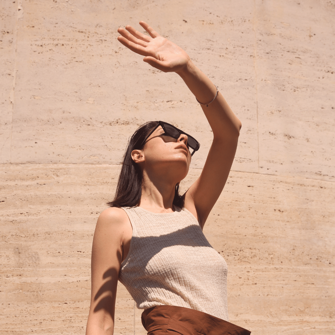 Une femme portant des lunettes de soleil et un chapeau, protégeant sa peau sensible du soleil avec son bras levé.